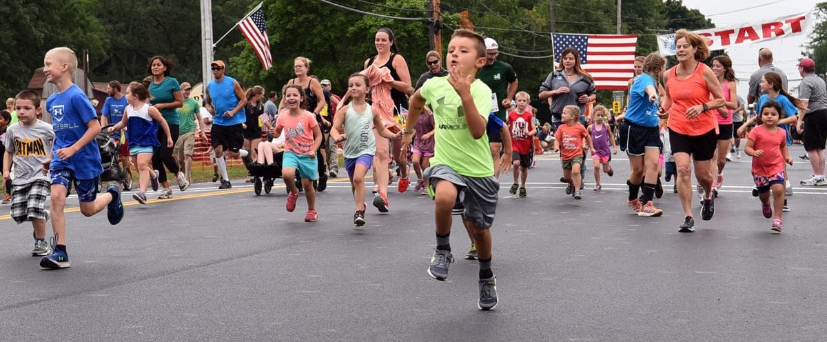 Children sprinting from the start line of the Great Race Kids' Fun Run, with American flags overhead.