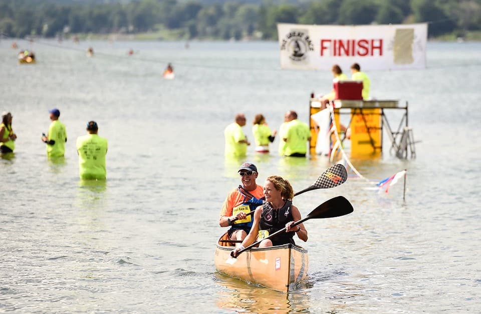 A team paddles a wooden canoe toward the Great Race FINISH banner on Owasco Lake while volunteers in neon shirts wait in the shallows.
